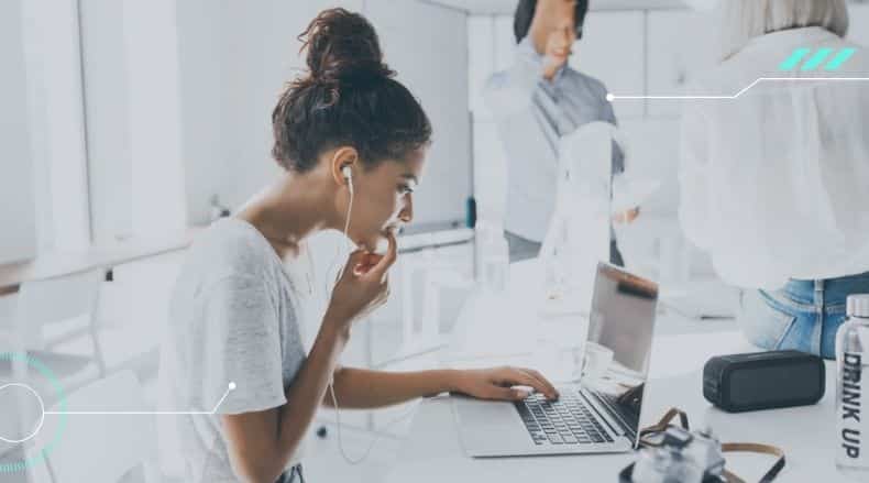Young woman with earbuds focused on laptop at a bright shared workspace with other people.