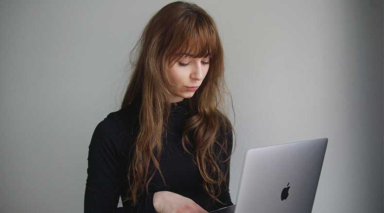 Young woman with long brown hair looking at a silver Apple laptop against a plain wall.