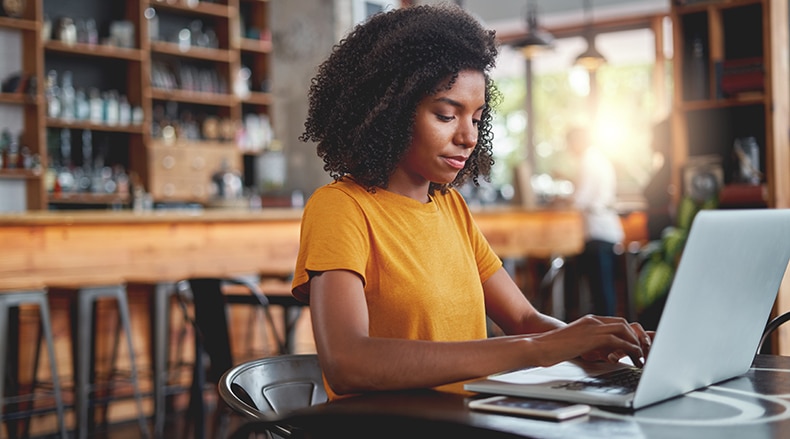 Woman in orange shirt working on a laptop in a cozy cafe with blurred background.