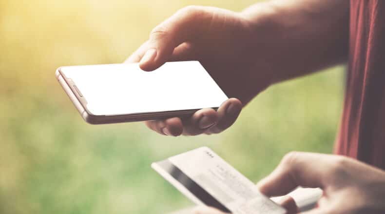 Person holding a smartphone and a credit card outdoors with blurred green background.