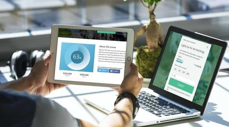 Person holding a tablet showing a fundraising progress chart with a laptop displaying an online form in the background.