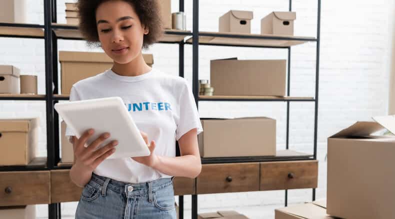 Woman wearing volunteer shirt looking at a tablet in a storage room with cardboard boxes on shelves.