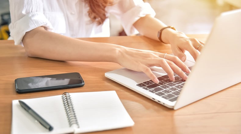 Person typing on a laptop with a phone and notebook with pen on wooden desk.