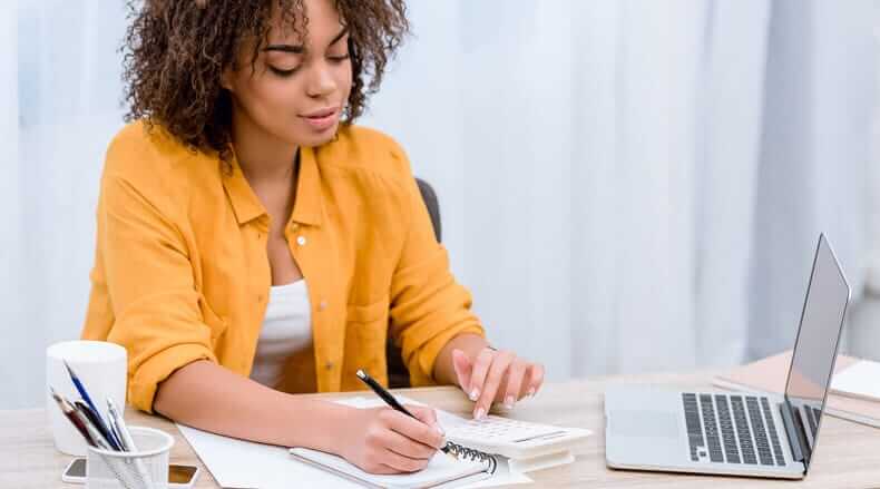 Woman in yellow shirt writing in a notebook with laptop and pens on the desk in front of her.