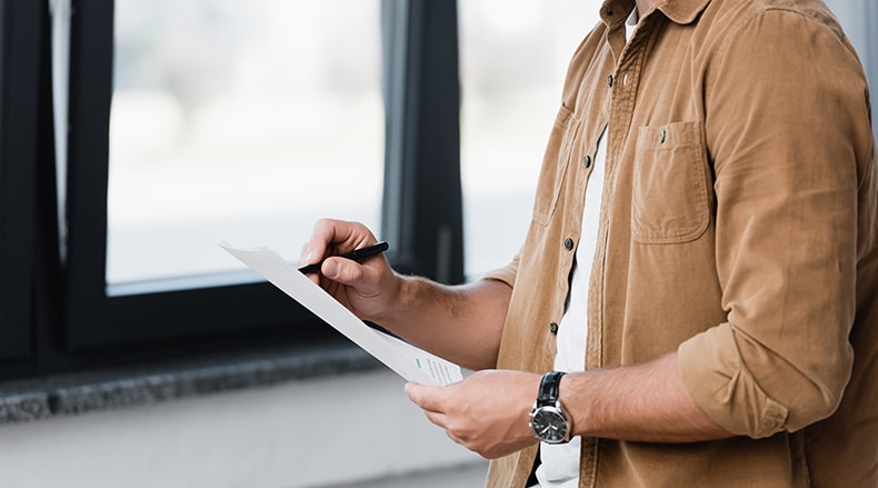 Person in brown shirt holding a pen and signing or writing on a document near a window.