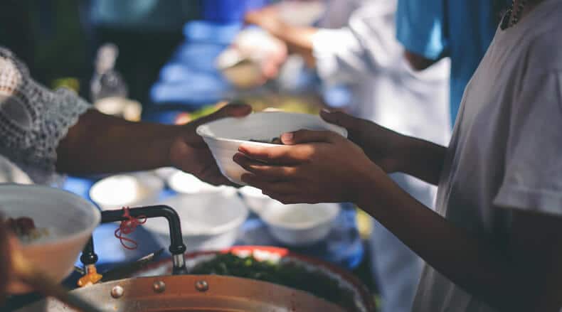 Two people exchanging a white bowl at a food serving station under warm lighting.