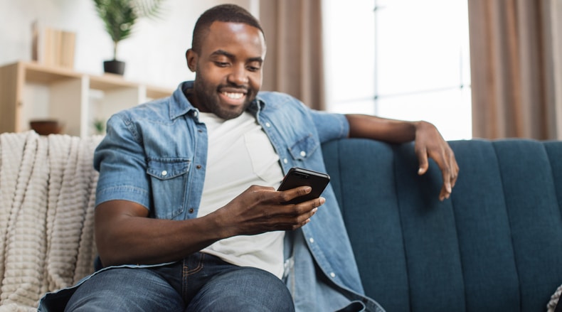Smiling man sitting on blue couch, looking at phone in hand in a bright room.