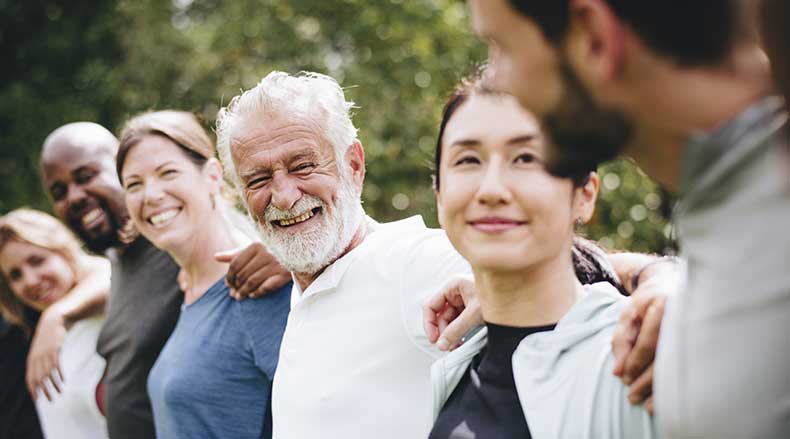 Group of diverse adults standing arm in arm outdoors, smiling and looking at each other.