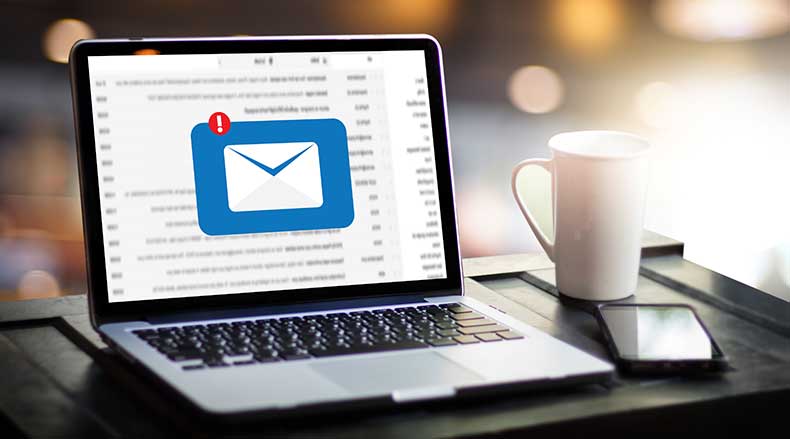 Open laptop on desk showing email inbox with new message notification, beside a coffee mug and smartphone.