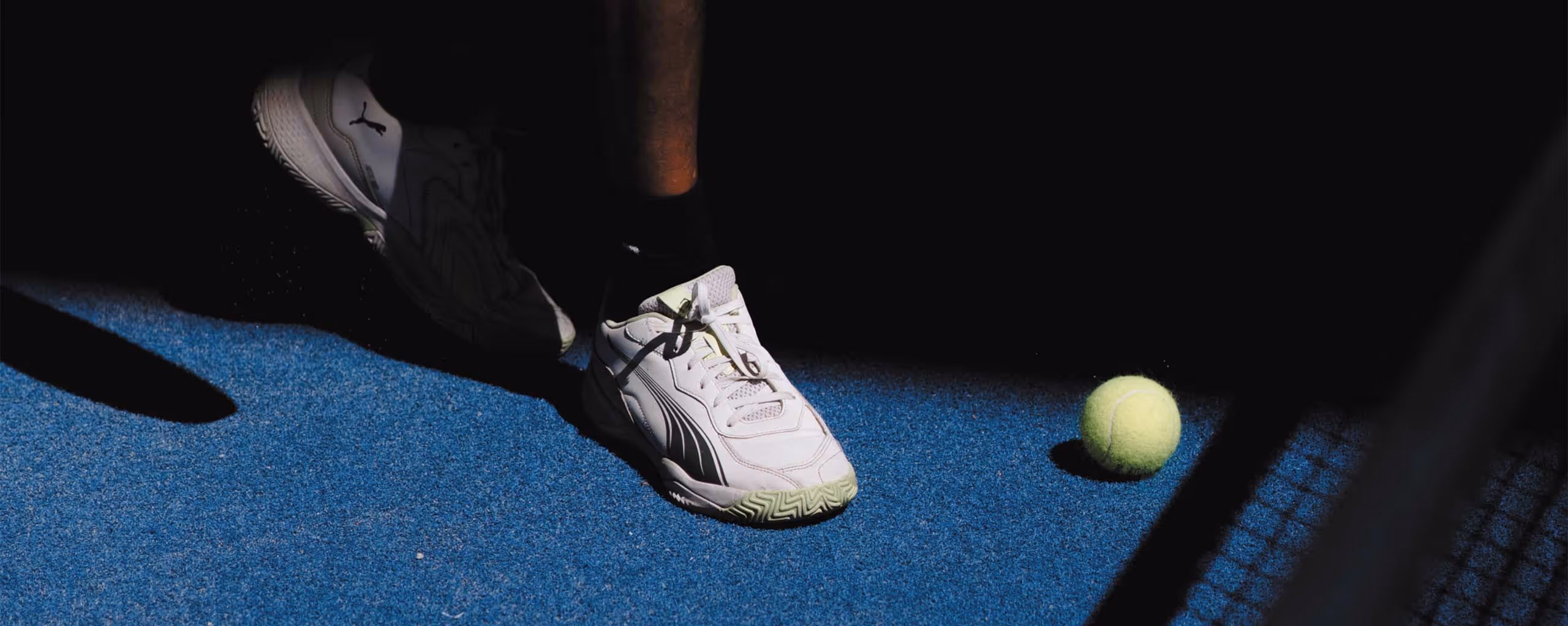 Primer plano de una persona usando tenis Puma blancas en una cancha azul, cerca de una pelota de pádel amarilla y una red de pádel.