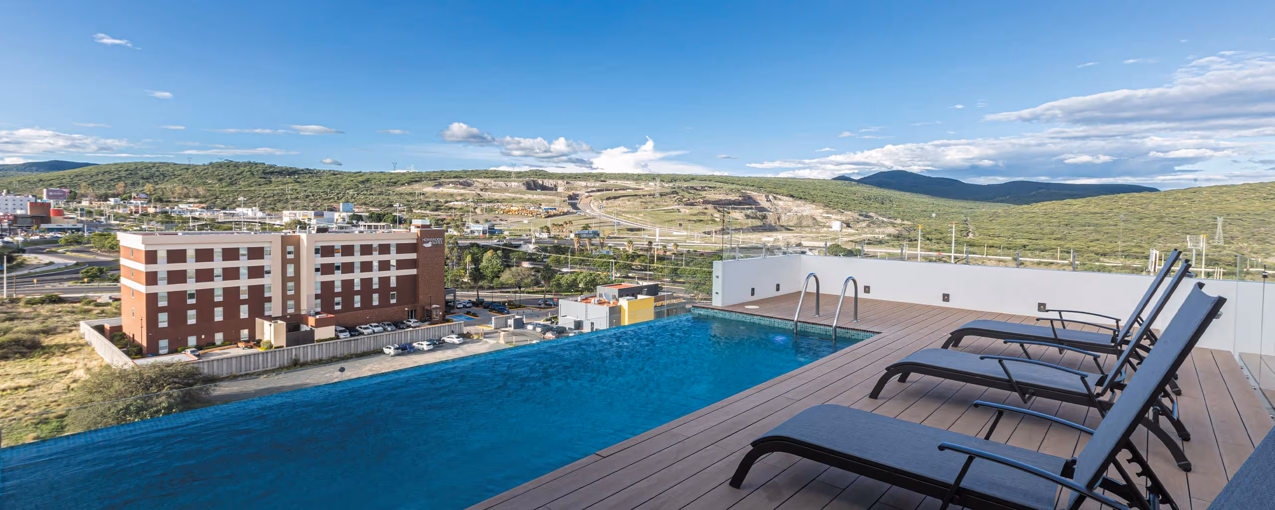 Alberca infinita en la azotea con cuatro camastros, vista a colinas verdes y un edificio hotelero.