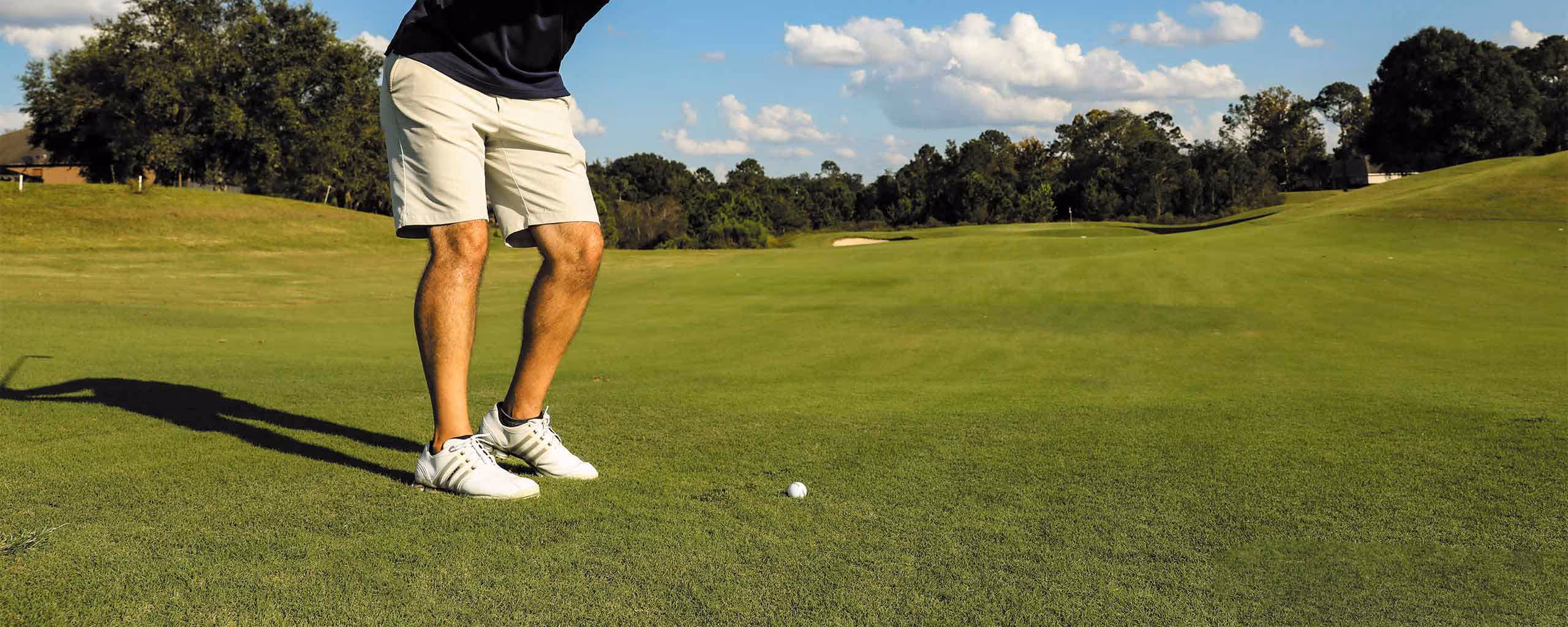 Piernas de un golfista con pantalones cortos y zapatos blancos preparado para golpear una bola de golf en un campo verde bajo un cielo azul con nubes.