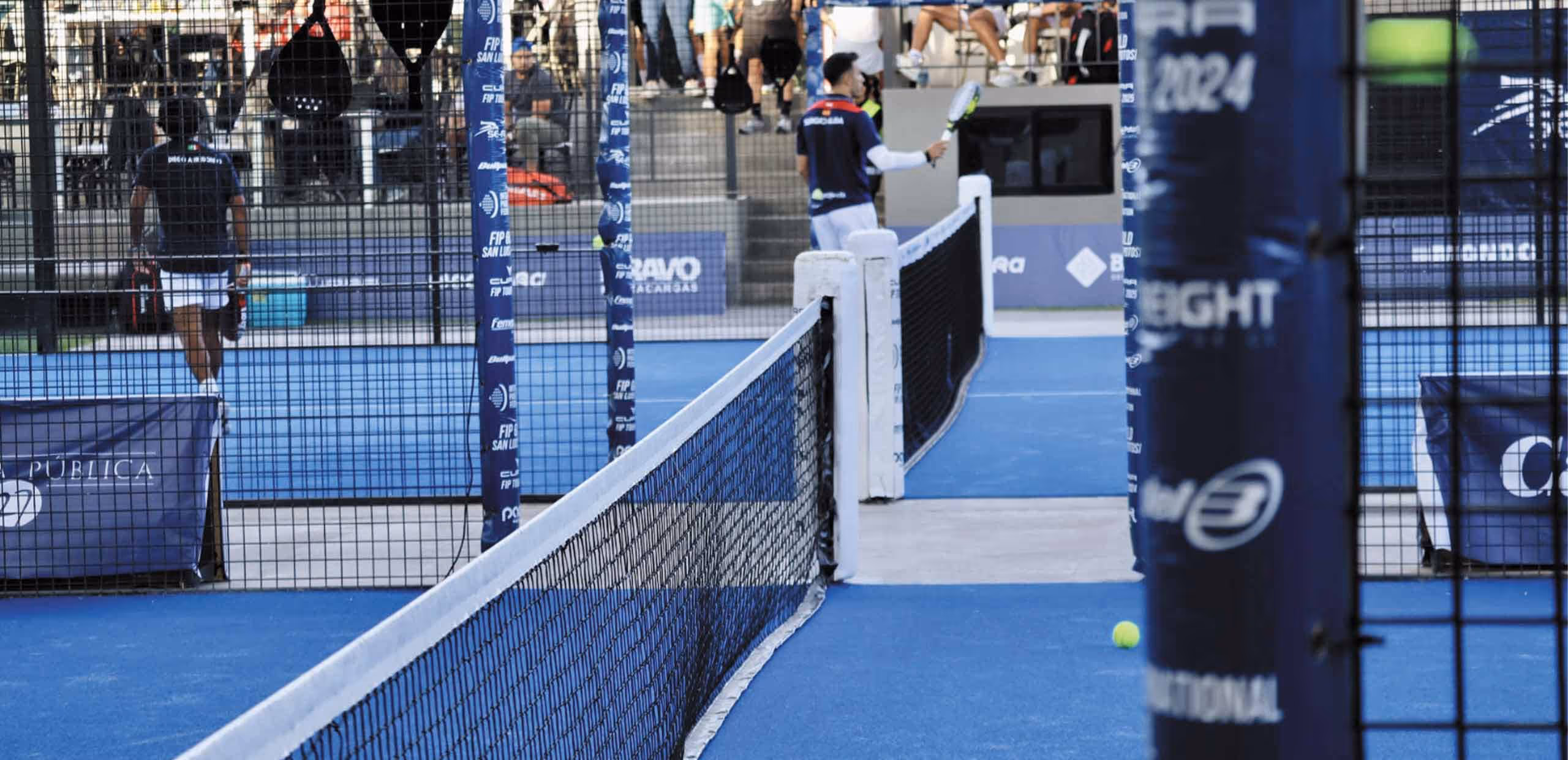 Dos jugadores en una cancha de pádel azul con red y público al fondo.