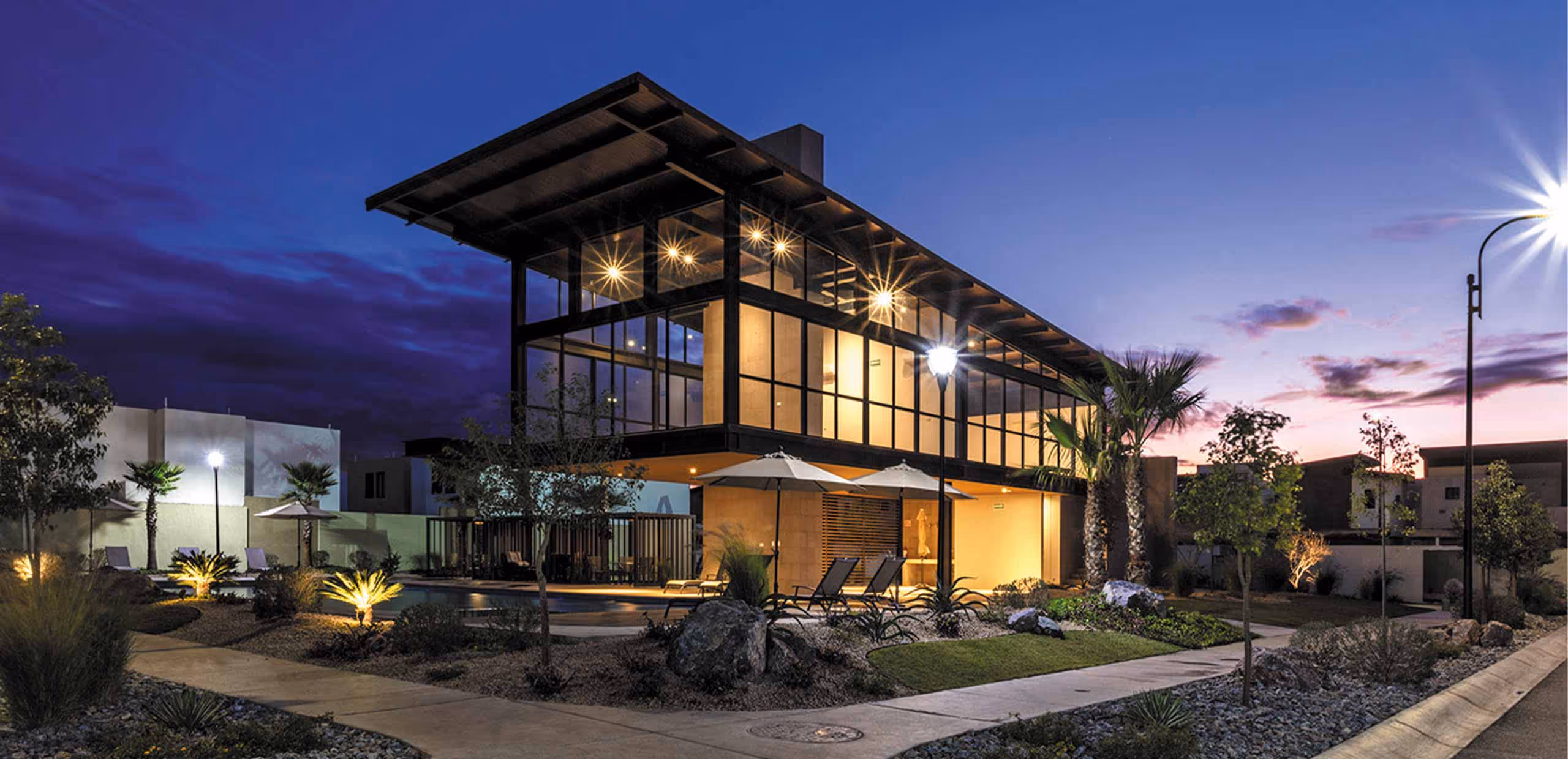 Casa moderna con ventanas grandes iluminada al atardecer, con jardín cuidado y palmeras.