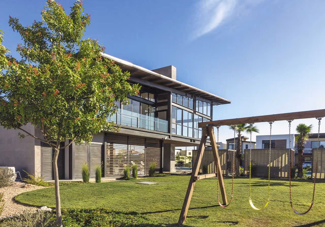 Casa moderna de dos pisos con grandes ventanales y un jardín con césped, un árbol y un columpio de madera con tres asientos amarillos.