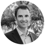 Smiling man in a suit standing outdoors with leafy trees in the background.