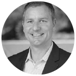 Smiling man in a suit and light shirt against a blurred background.