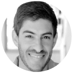 Smiling man with short dark hair and light facial hair in a blurred indoor setting.