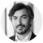 Black-and-white headshot of a man with dark hair and beard, wearing a white shirt and dark jacket.