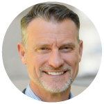 Smiling middle-aged man with short gray hair and a light beard, wearing a blue shirt.