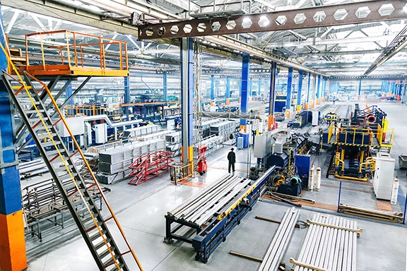 Spacious industrial factory floor with machinery, metal pipes, storage racks, and a worker in the middle.