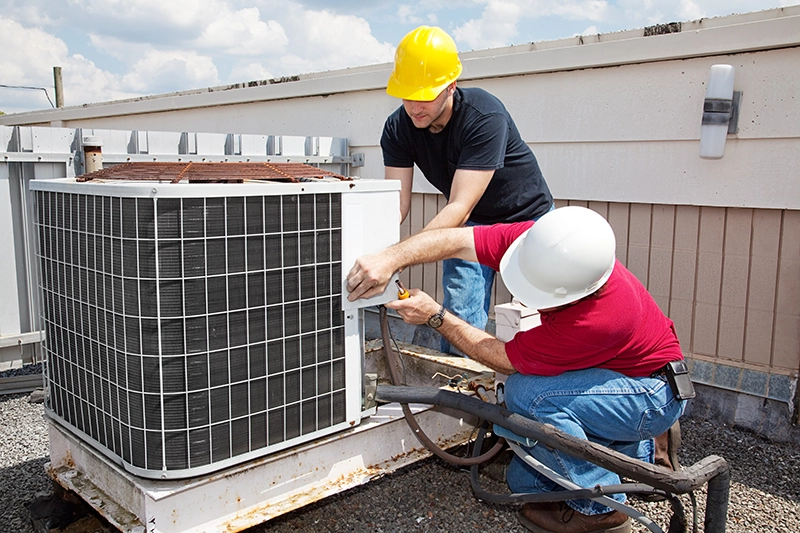 Two HVAC technicians wearing safety helmets repairing an outdoor air conditioning unit on a rooftop.