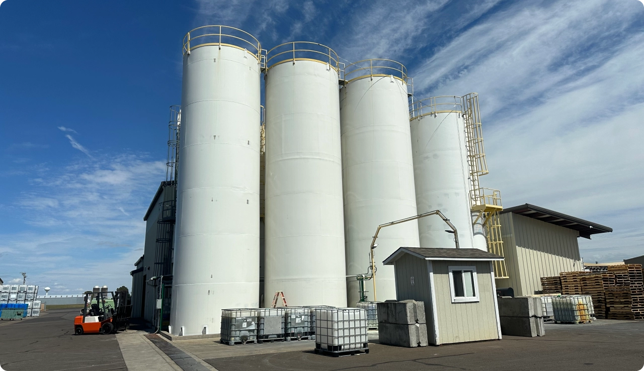 Four tall white industrial silos with yellow railings and ladders next to a small gray guardhouse and stacks of pallets under a partly cloudy blue sky.