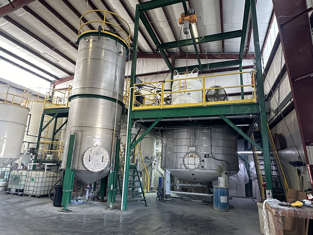 Industrial facility interior with large stainless steel tanks and green metal framework, pipes, and yellow safety railings.