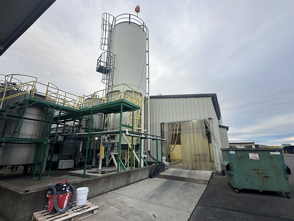 Industrial facility exterior with large white vertical tank, green metal framework, beige building with plastic strip curtains, and a green dumpster on a cloudy day.