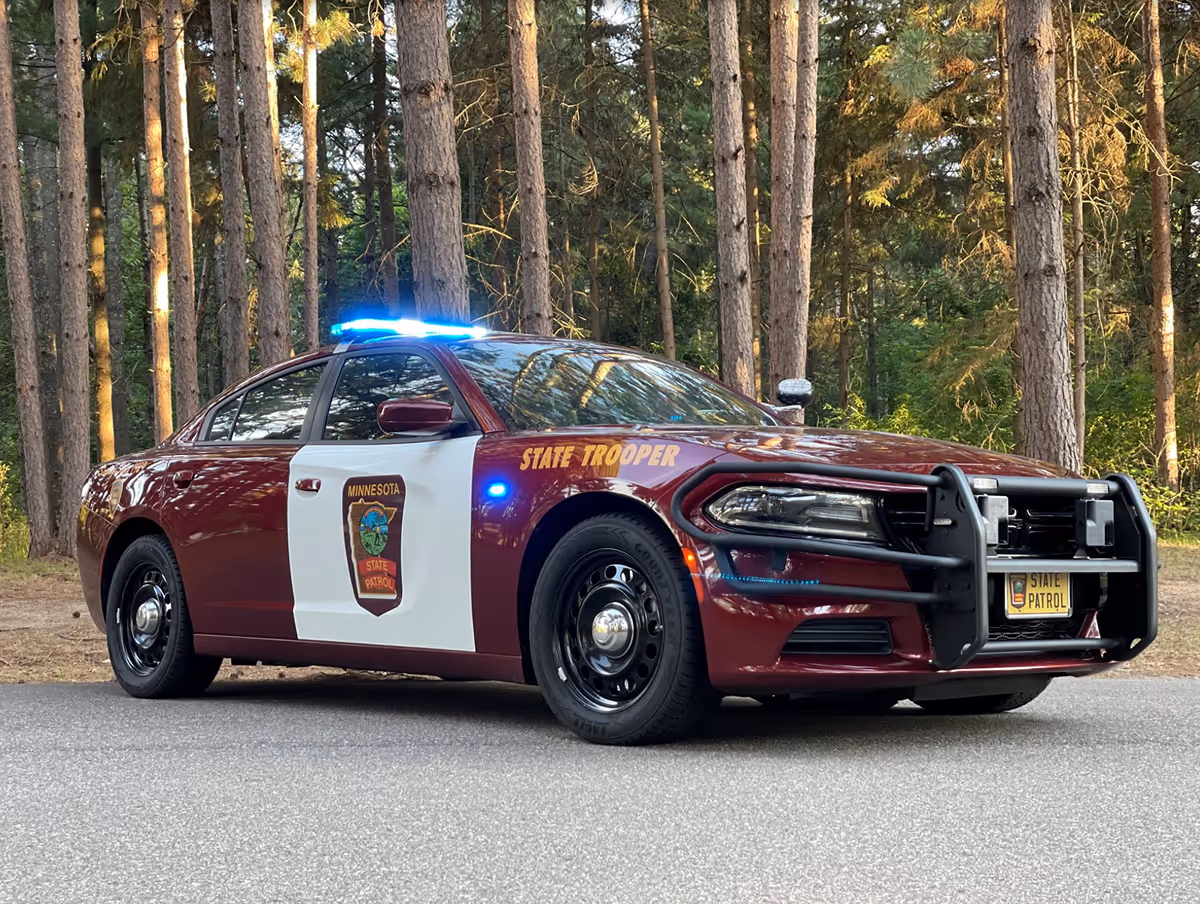 Maroon and white Minnesota State Patrol police car with blue lights on, parked on a road with tall trees in the background.