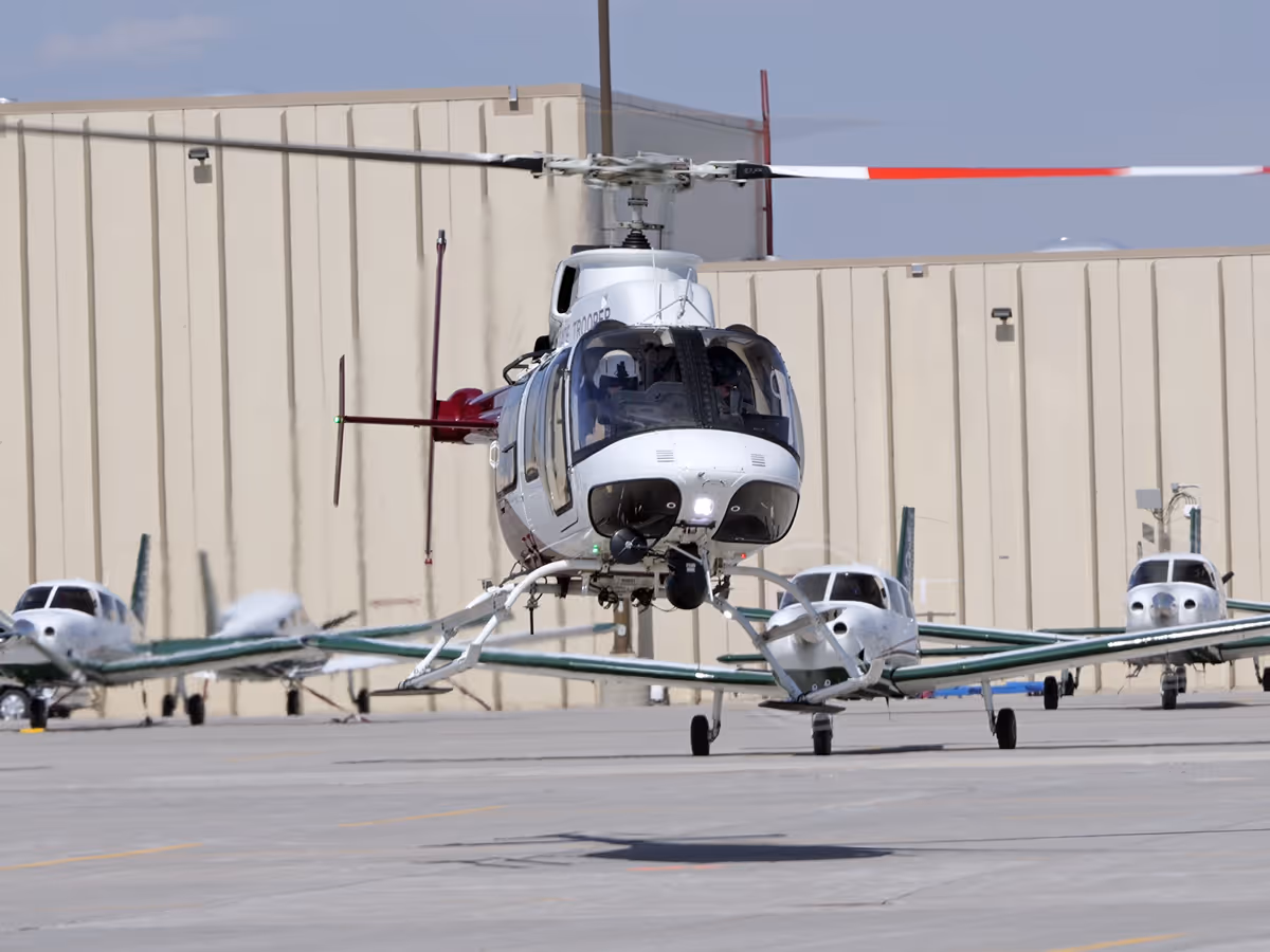 White and gray helicopter hovering above the tarmac with small propeller airplanes parked in the background.