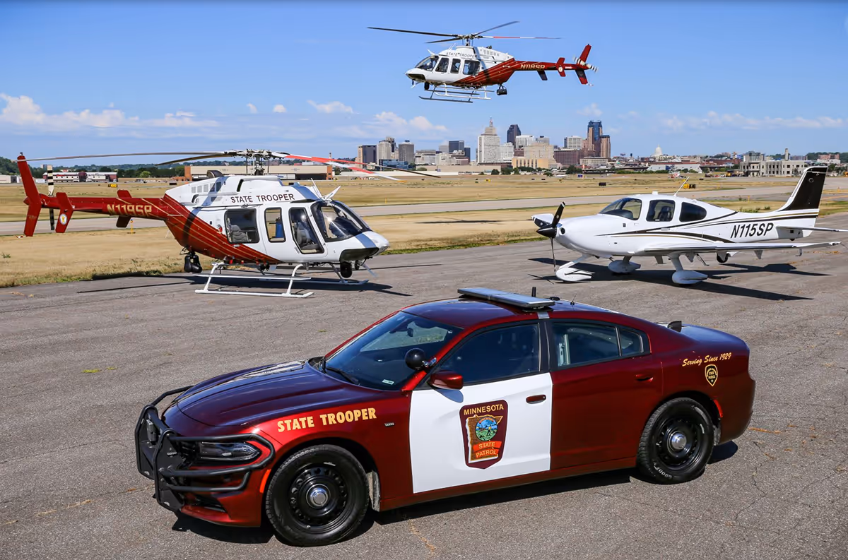 Minnesota State Patrol maroon and white car parked on tarmac near a white single-engine plane and two red and white state trooper helicopters, with a city skyline in the background.