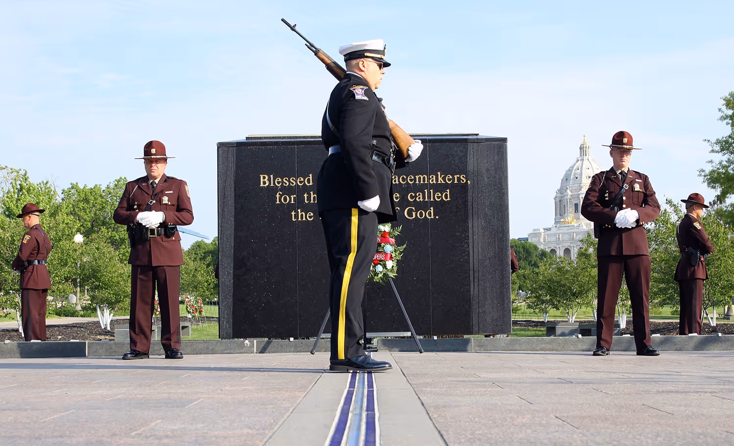 Military honor guard standing at attention in front of a black memorial stone with inscription about peacemakers.
