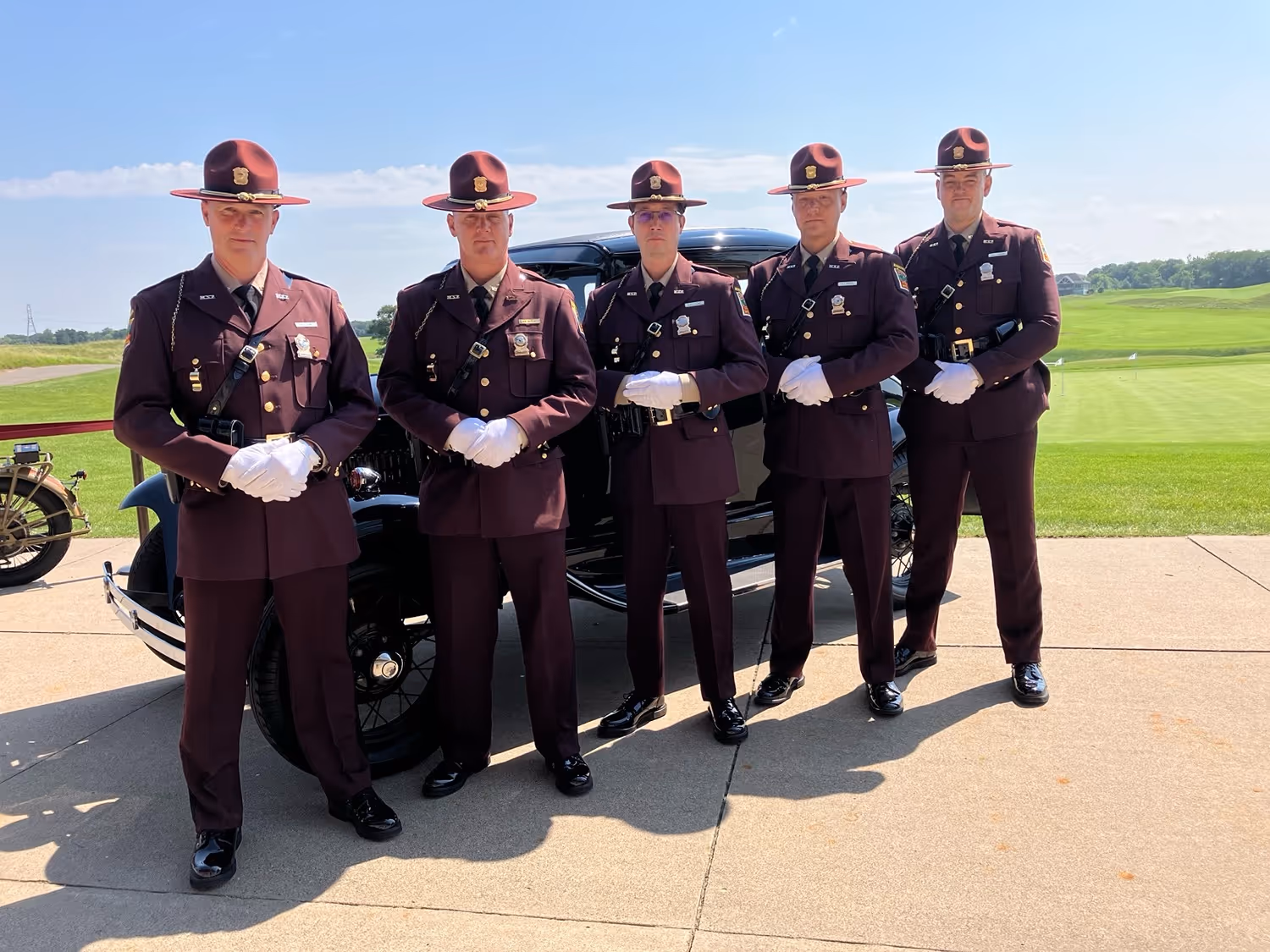 Five uniformed police officers wearing maroon hats and white gloves standing in front of a vintage black car on a sunny day.