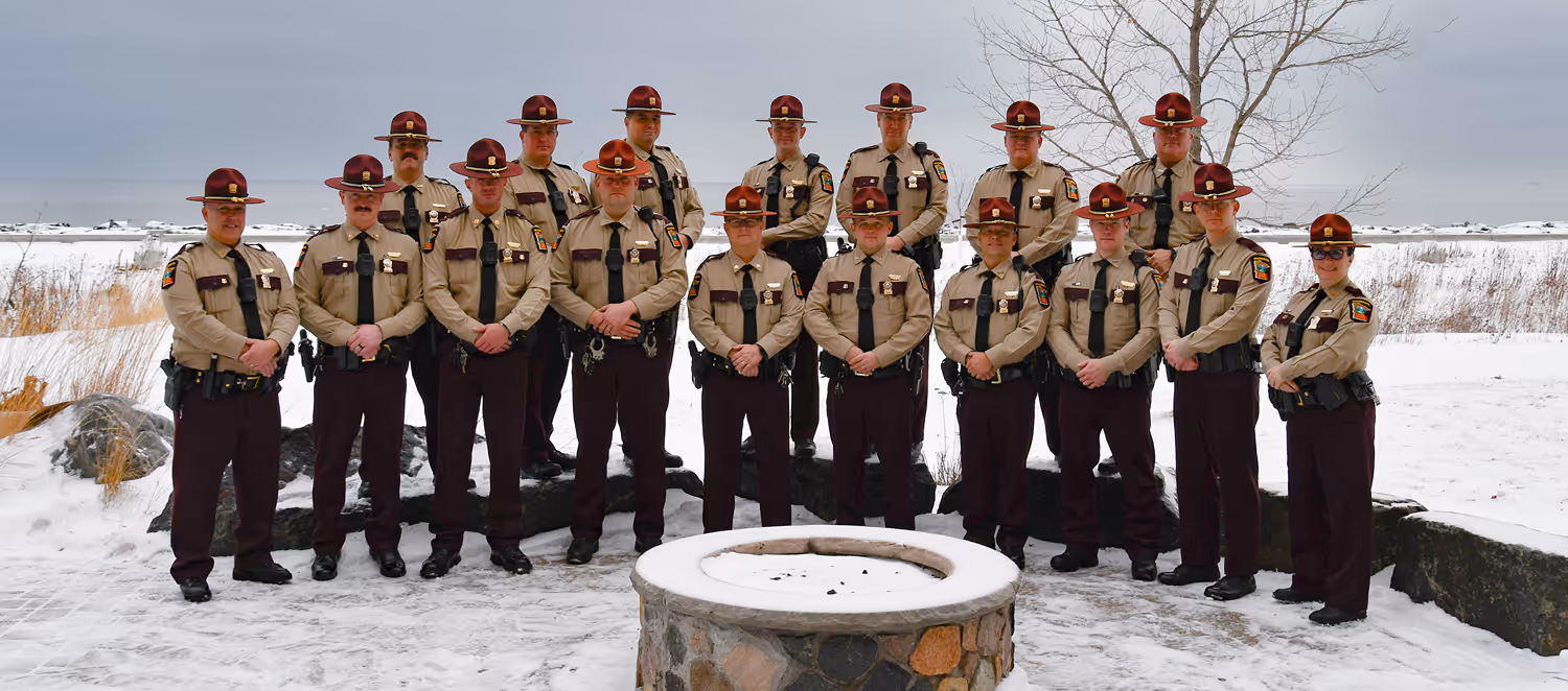 Group of sixteen uniformed law enforcement officers standing on snowy ground near a stone fire pit with a winter landscape in the background.