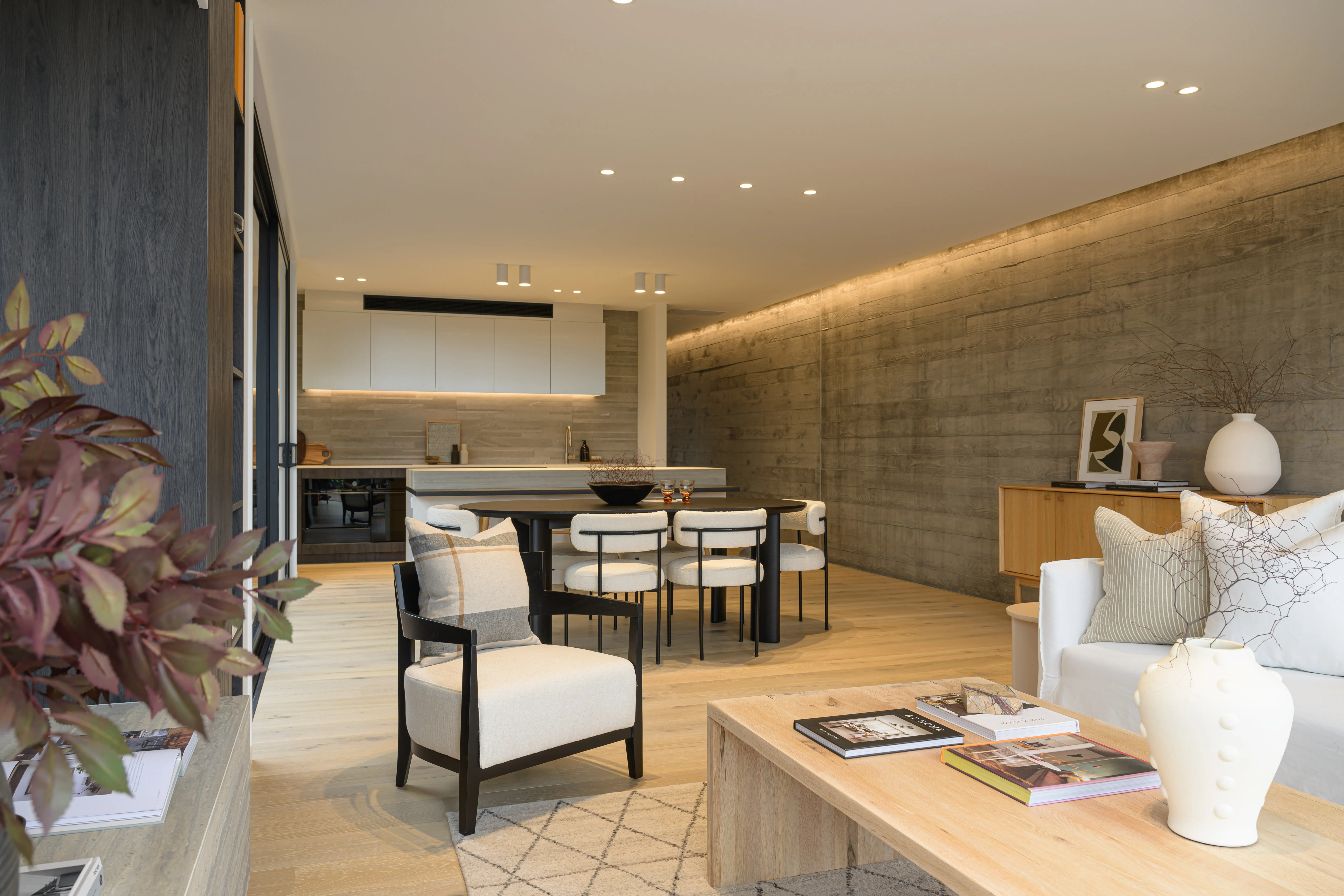 Modern open living room and kitchen with neutral decor, beige armchair, white sofa, wooden table, and dining area.