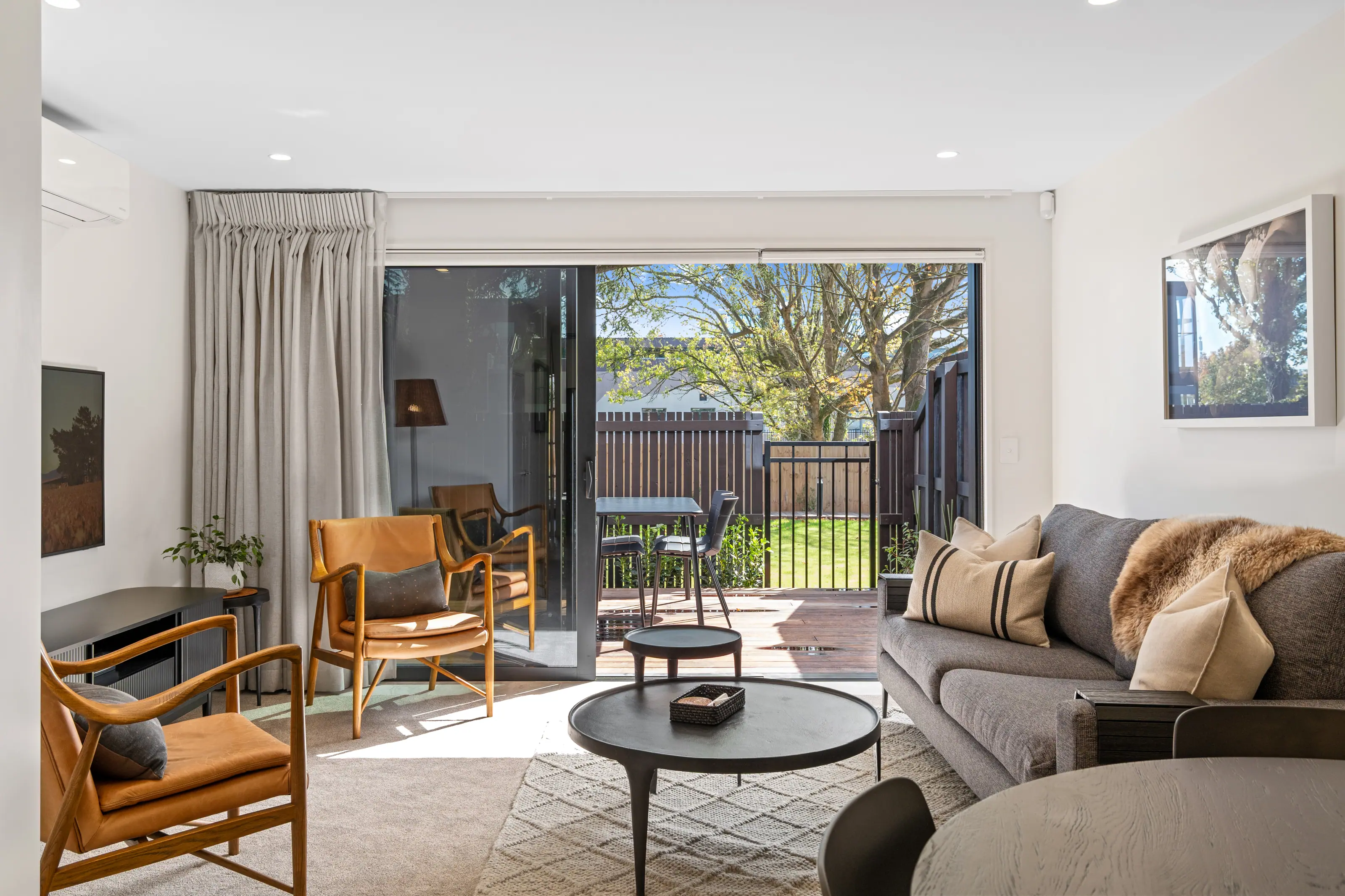 Modern living room with grey sofa, two leather chairs, round coffee table, and sliding glass door opening to outdoor patio.
