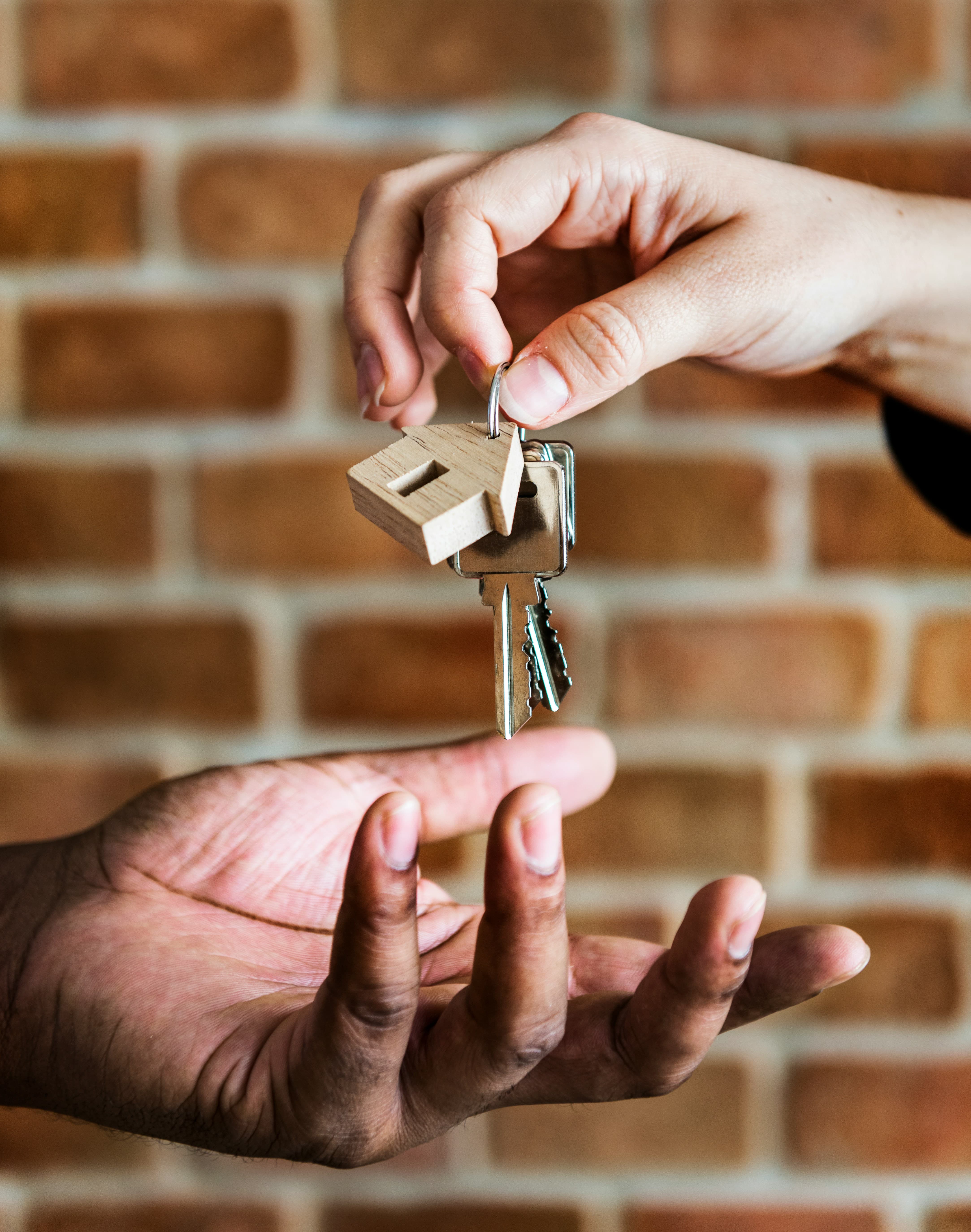 Hand holding house-shaped keychain with keys, extending keys to an open palm, brick wall background.