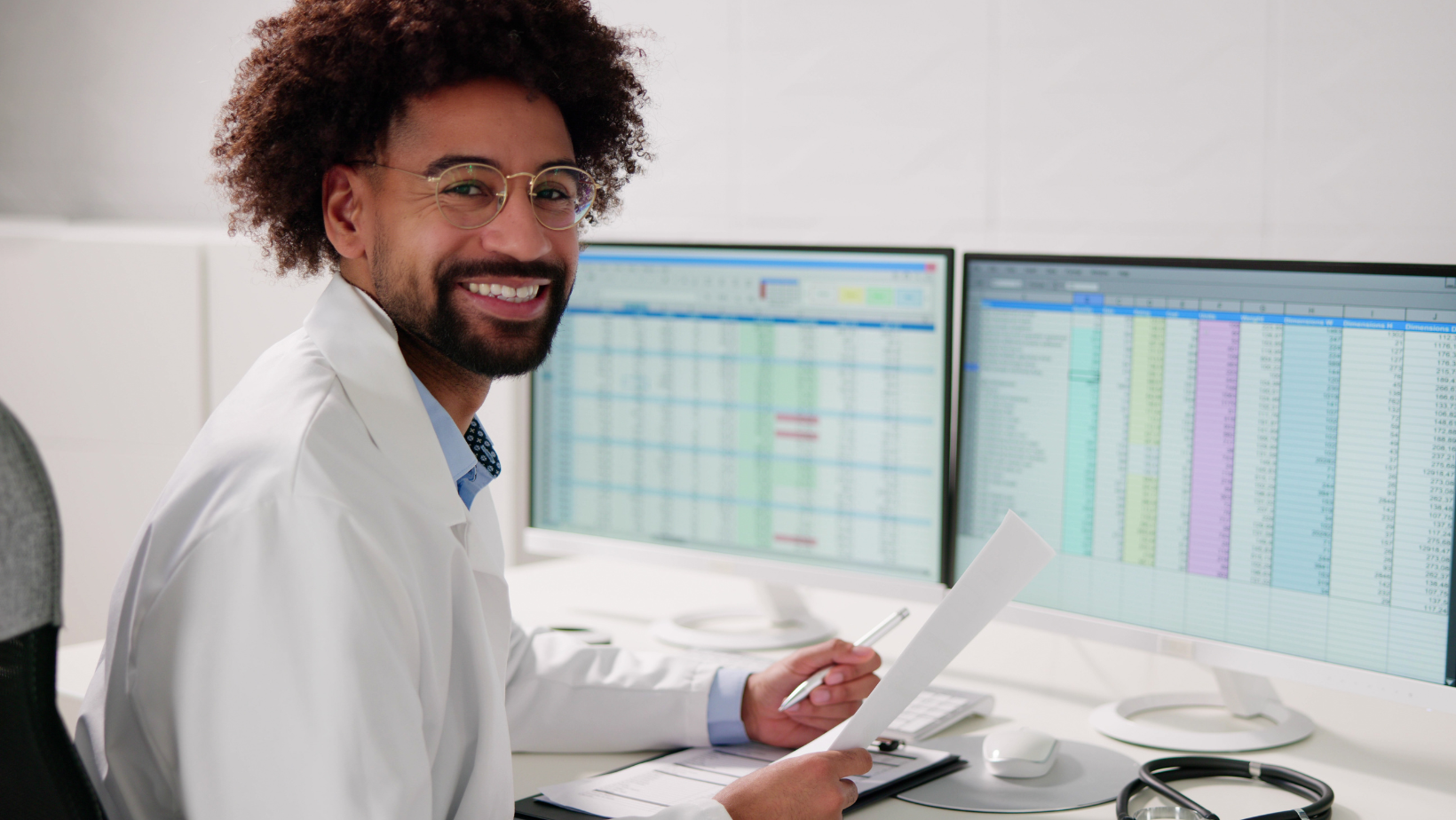 Smiling doctor in a white coat holding a paper and pen, sitting at a desk with two monitors displaying spreadsheets.