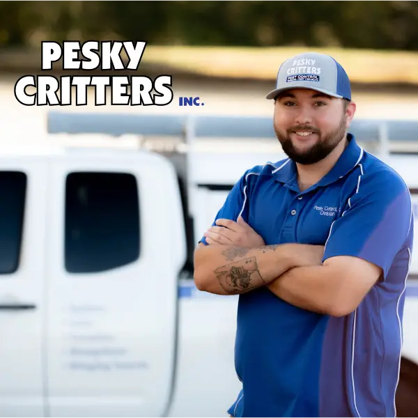 Pesky Critters Inc. pest control technician in blue uniform and cap standing with arms crossed in front of company truck.