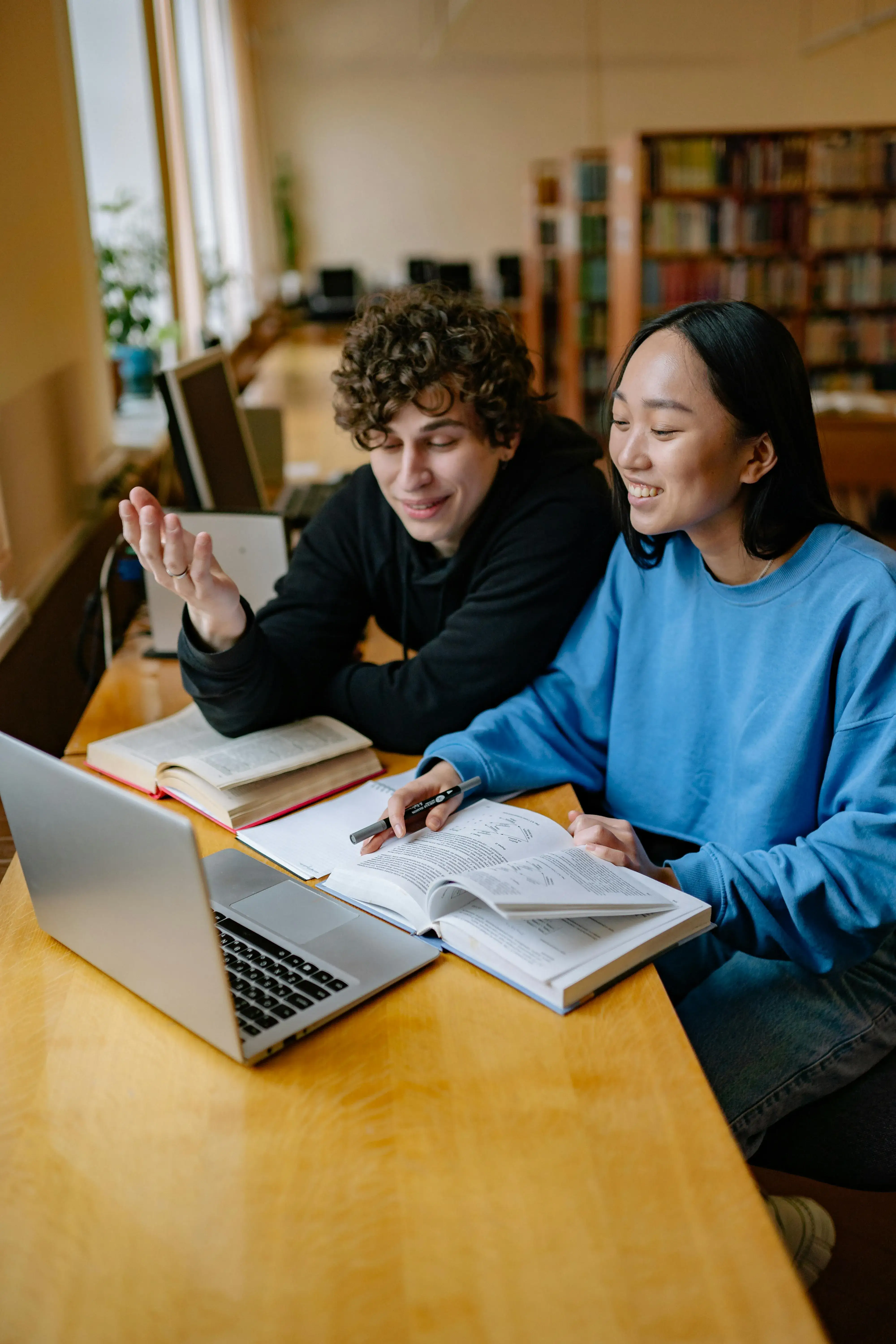 Two students sitting at a wooden table in a library, smiling and studying together with open books and a laptop.