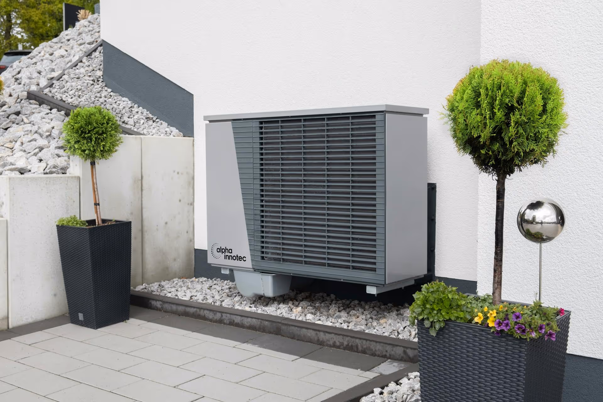 Modern gray and white outdoor heat pump unit mounted on a white wall between two potted topiary plants with flowers.