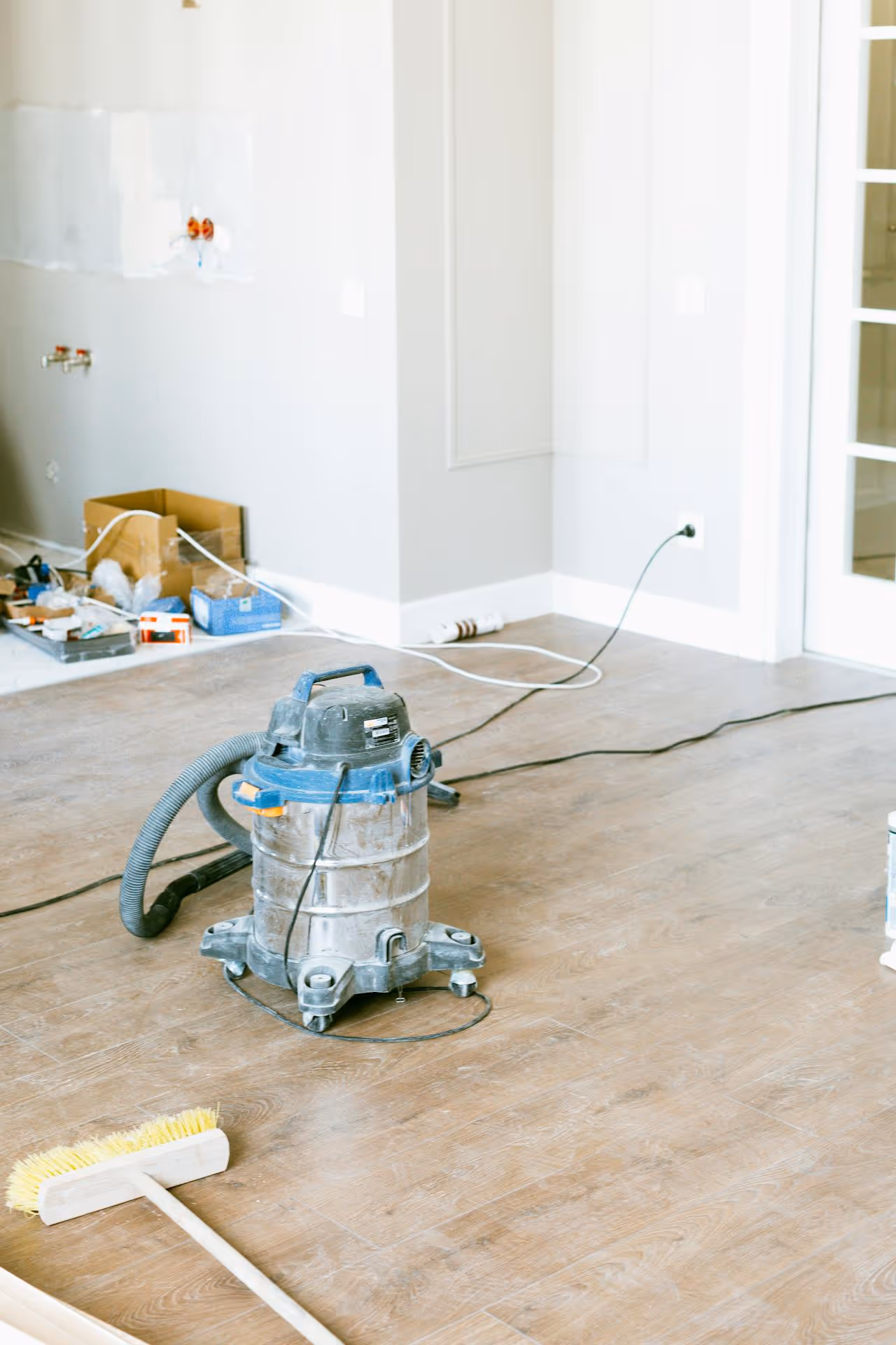 Wet/dry vacuum cleaner on a hardwood floor in a partially renovated room with a broom and scattered tools in the background.