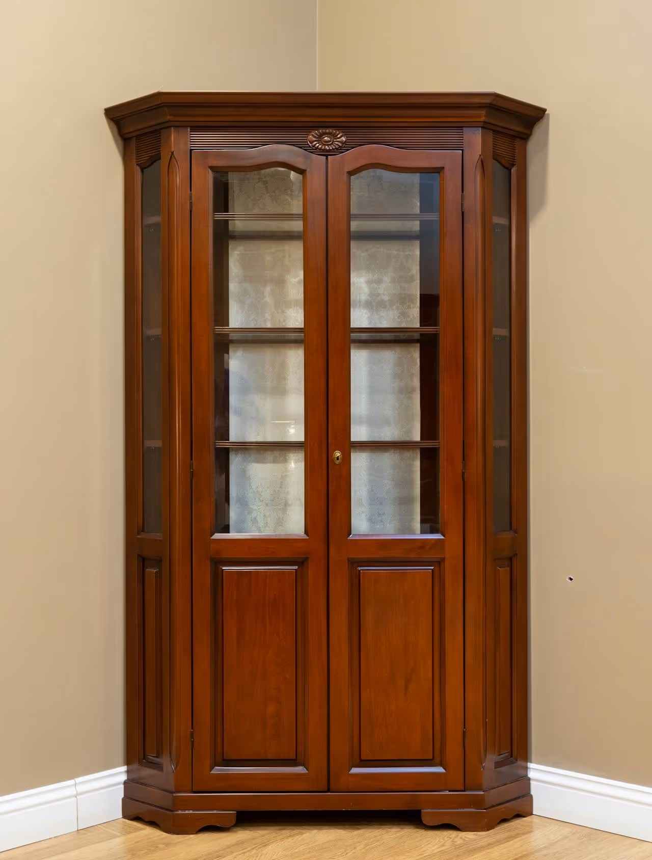 Wooden corner cabinet with glass doors and shelves inside, standing on a hardwood floor against beige walls.