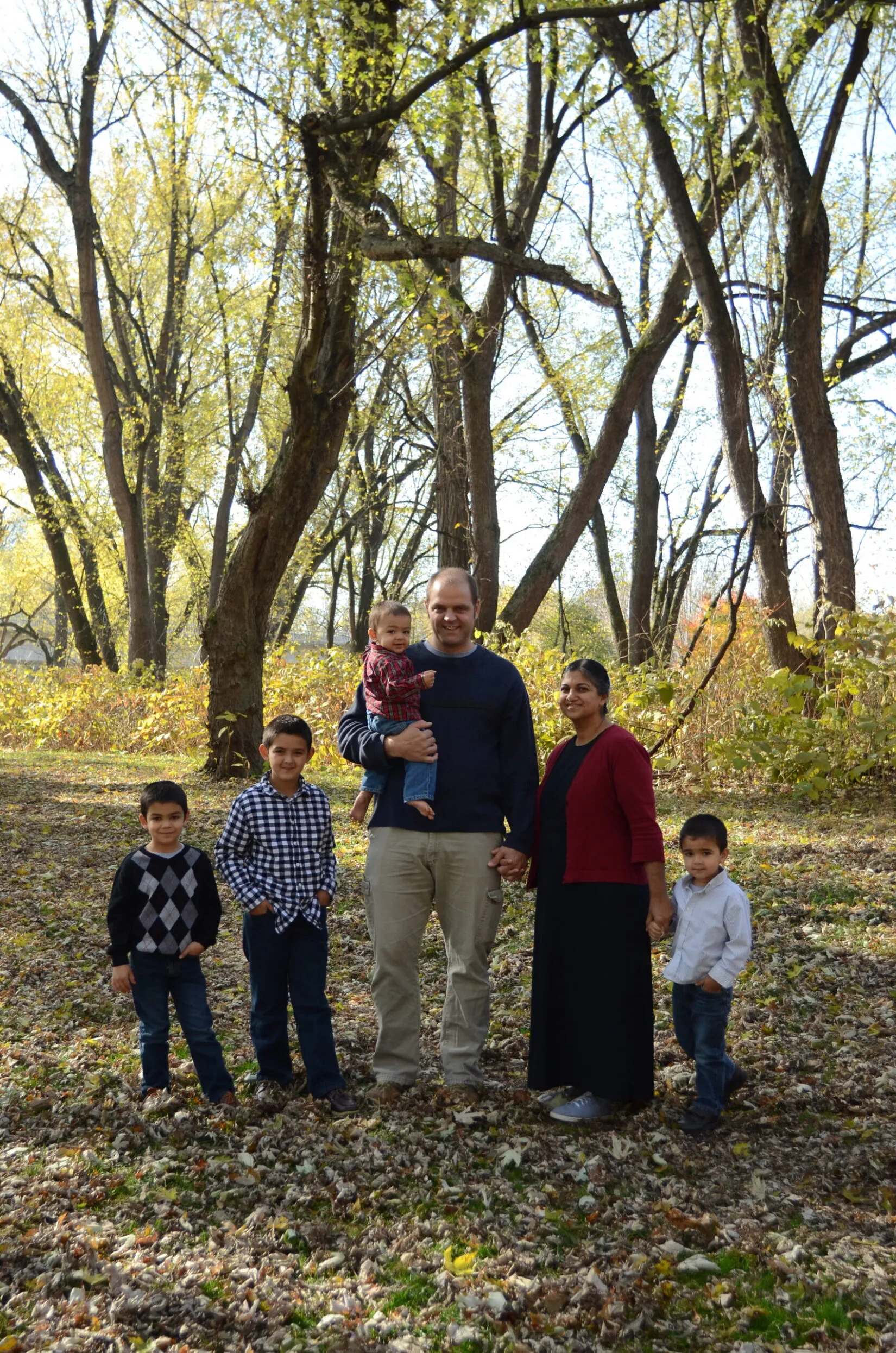 Family of six standing outdoors on a leafy ground with tall trees in the background, smiling and holding hands.