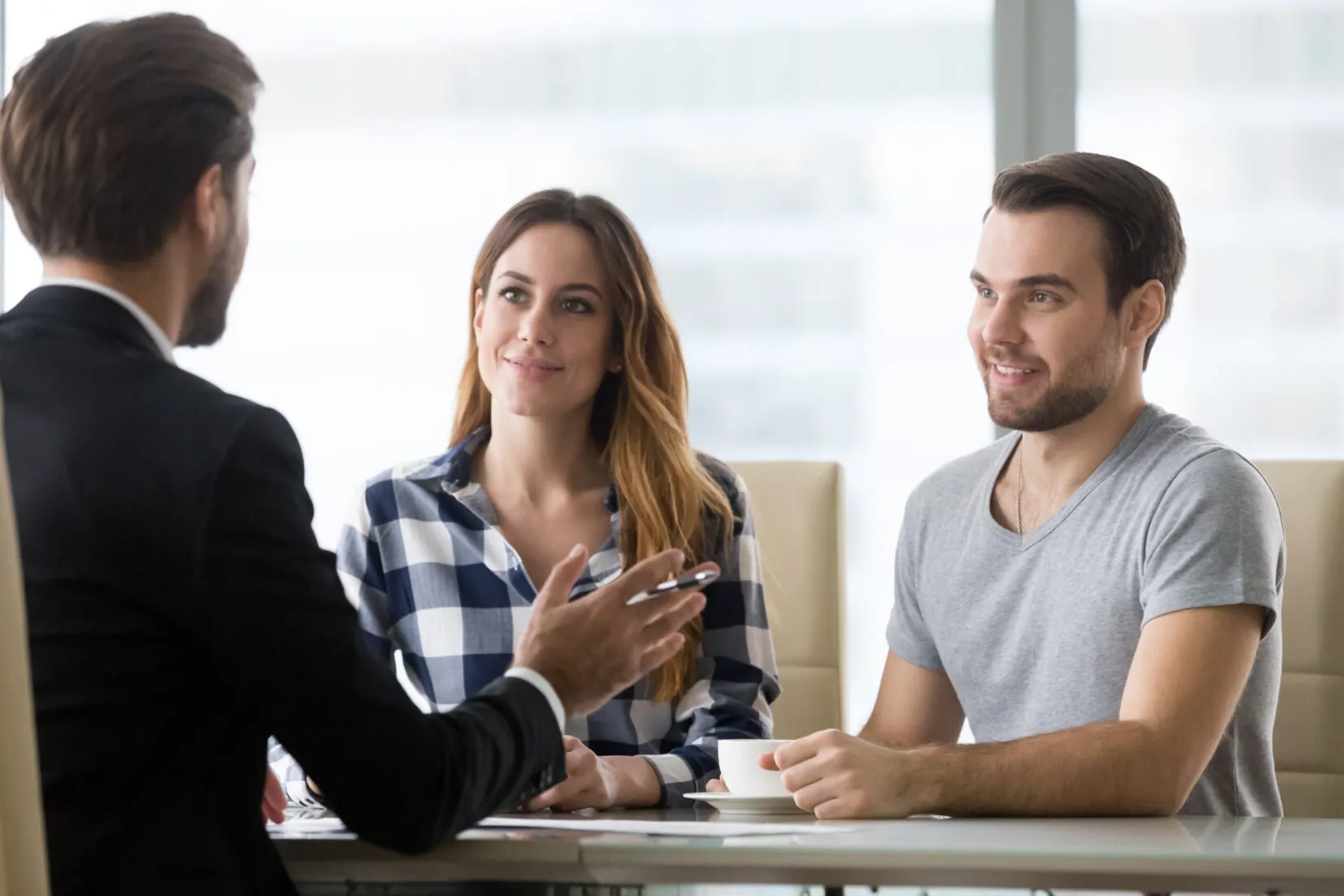 A man in a suit talks to a smiling couple sitting at a table with a cup of coffee.