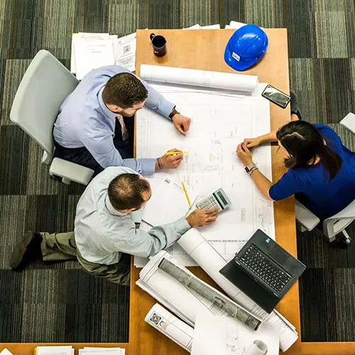 Three professionals collaborating over architectural blueprints at a wooden table with a laptop, calculator, and a blue safety helmet.
