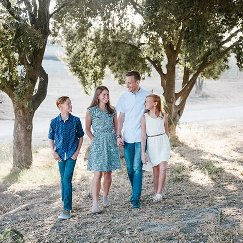 A family of four, including two parents and two children, walking together outdoors among trees on a sunny day.