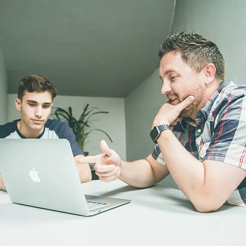Two men sitting at a table looking at a laptop, one pointing at the screen, in a bright room.