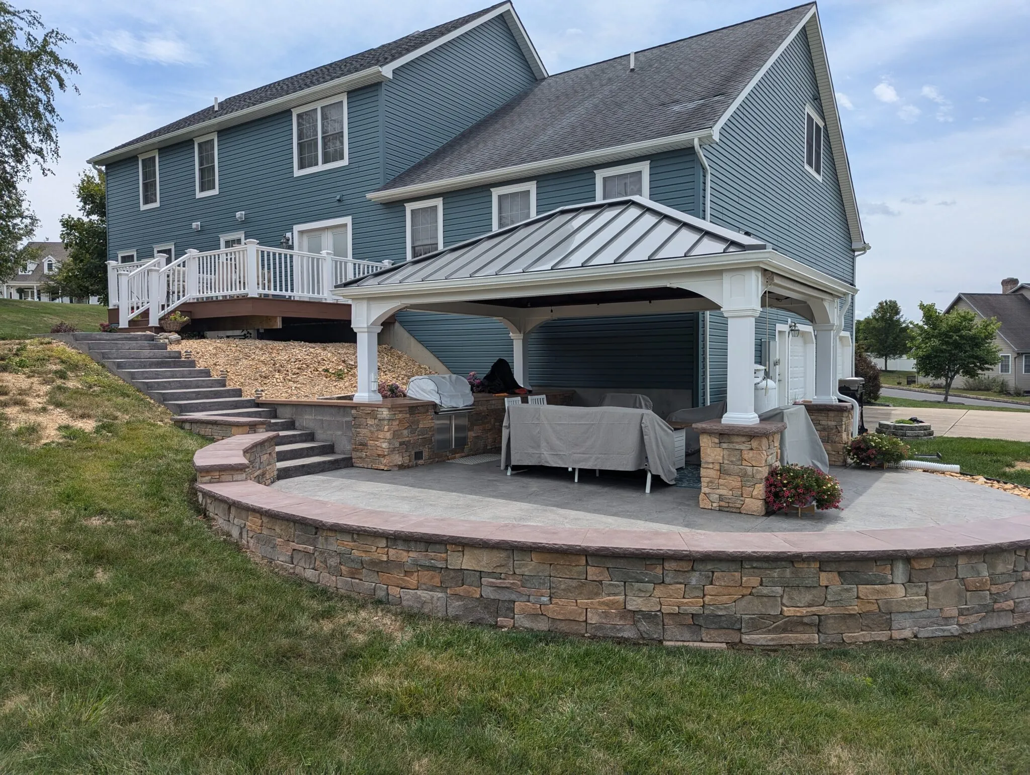 Covered outdoor patio area with stone pillars and seating covered in protective covers next to a blue two-story house.